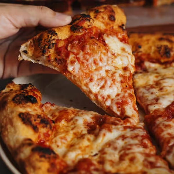 Hand lifting a slice of Crab Rock PIZZA with melted cheese and charred crust from a whole pizza.